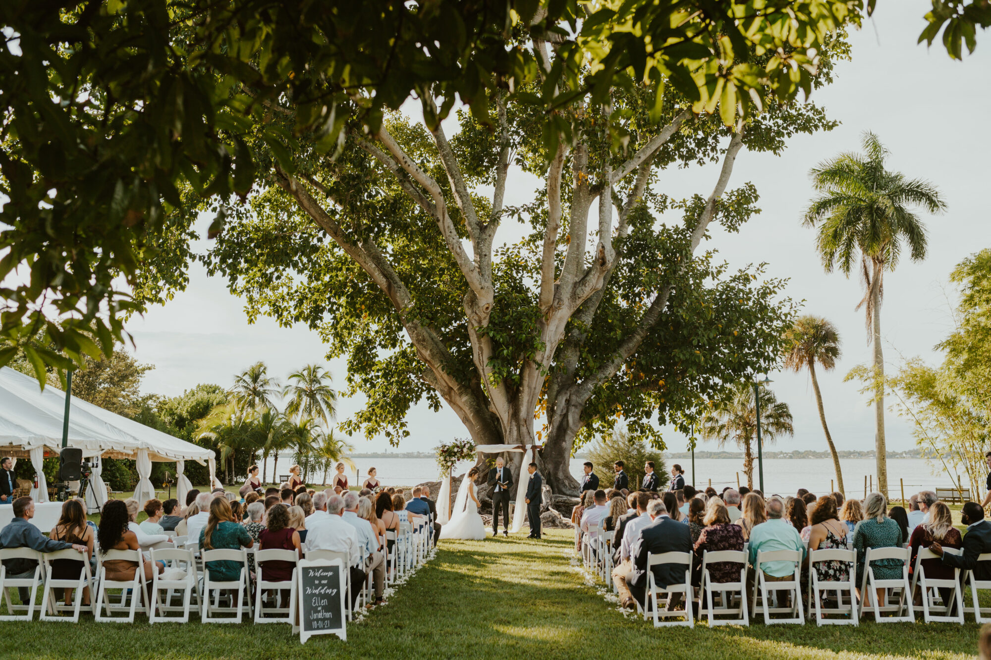 An outdoor wedding ceremony in progress at Edison and Ford Winter Estates, with guests seated under a large tree beside the river