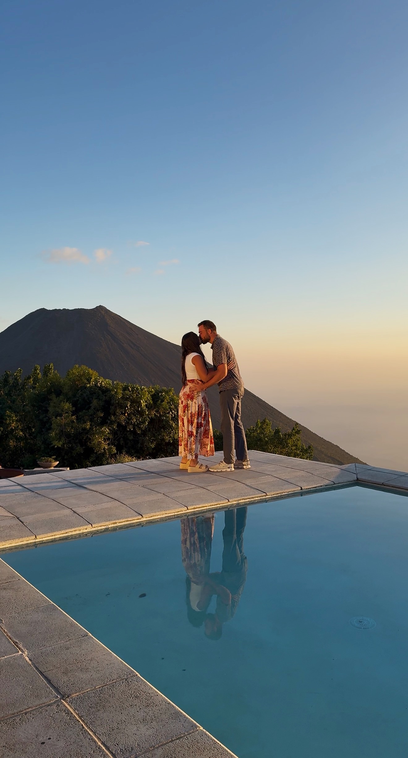 Evan and Corrina sharing a kiss at sunset with a volcano reflected in the infinity pool
