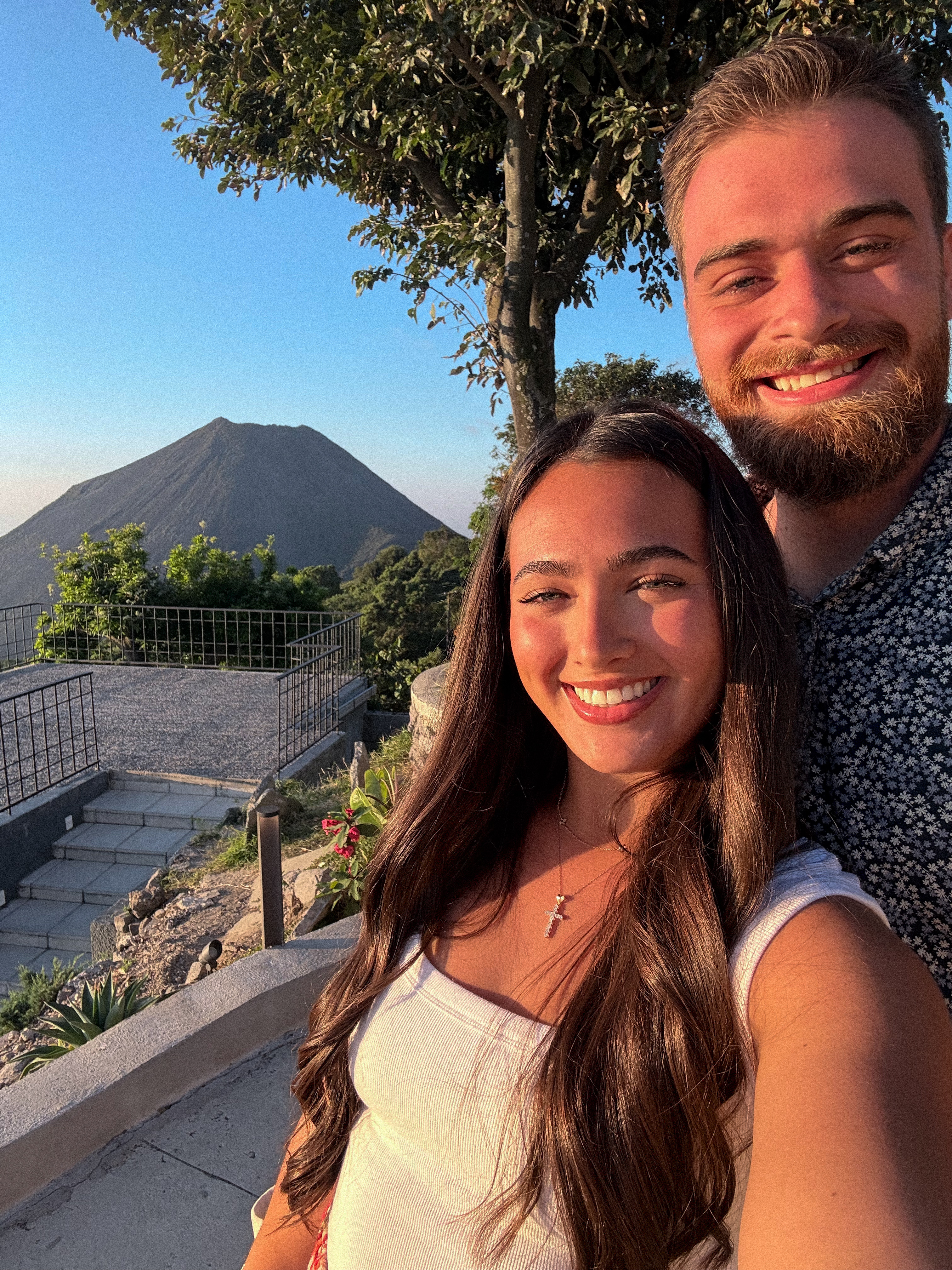 Evan and Corrina smiling together in a selfie with a volcano and golden sunset behind them